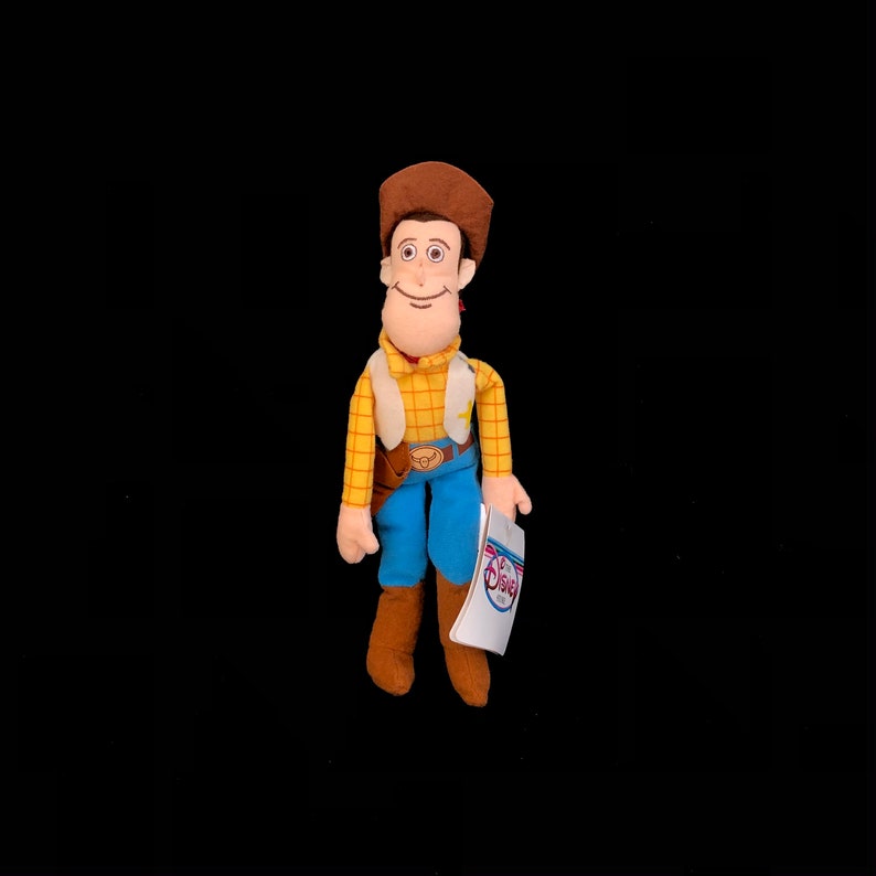 May include: A plush toy of the character Woody from the Disney Pixar film Toy Story. Woody is wearing a brown cowboy hat, a yellow plaid shirt, a tan vest, blue pants, and brown boots. He is holding a tag with the Disney logo.