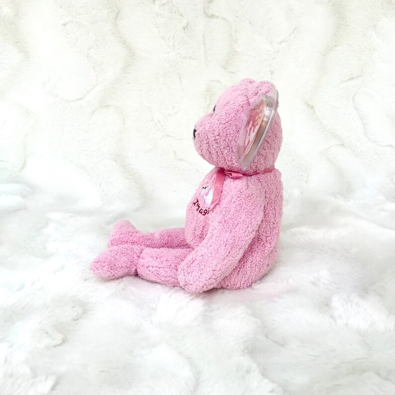 May include: Pink plush beanie baby with a white ribbon and the word "Angel" embroidered on its chest. The bear is sitting on a white fluffy surface.