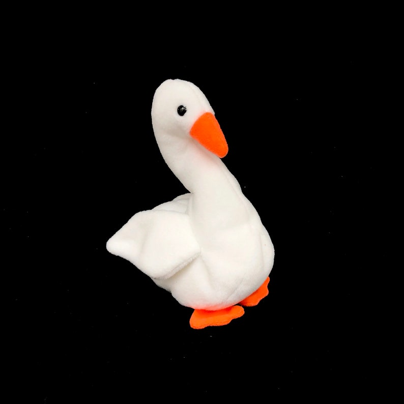 Op de afbeelding: Een witte pluche gans met een oranje snavel en poten. De gans heeft een gebogen nek en staat tegen een zwarte achtergrond. Het speelgoed is gemaakt van zacht, pluizig materiaal.