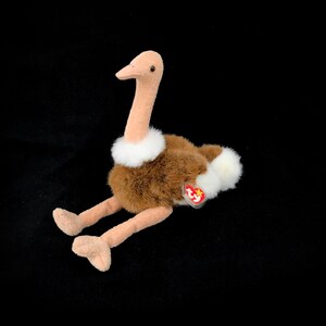 Puede incluir: Un juguete de peluche de avestruz con un largo cuello y pico color beige, cuerpo marrón y detalles blancos. El juguete tiene una etiqueta roja y blanca adjunta. La avestruz está sobre una superficie negra.