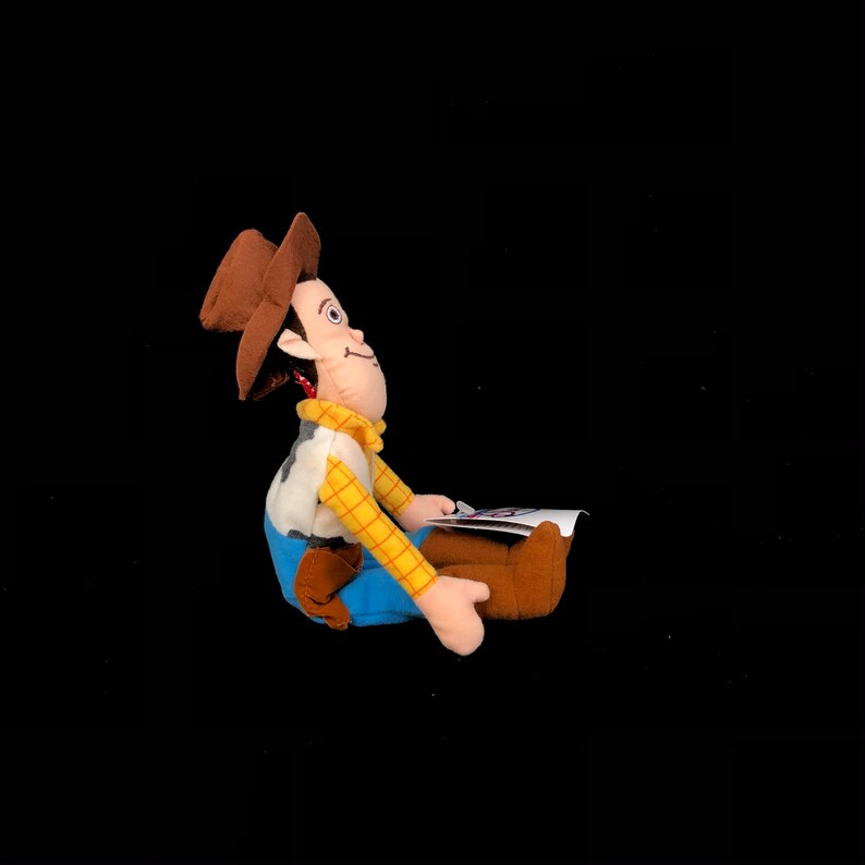 May include: A plush toy of a cowboy character, featuring a brown hat, a yellow plaid shirt, a white vest with a cow print, blue pants, and brown boots. The toy is sitting and holding a small card. The toy is a character from the Toy Story movie.