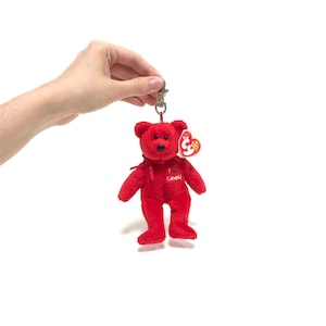 May include: A red plush bear keyring with a metal clasp. The bear has a small red bow and the words "I ❤️ Canada" embroidered on its chest. A small tag is attached to the bear's ear.