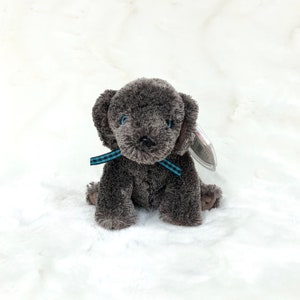 Puede incluir: Un perro de peluche gris con ojos azules y una cinta a cuadros azul y blanca alrededor del cuello. El perro está sentado sobre una superficie blanca y esponjosa.