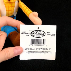 May include: A tag from The Disney Store for a Mini Bean Bag Woody doll, approximately 9 inches in height. The tag features the Disney Store logo and barcode, with text indicating exclusive availability.