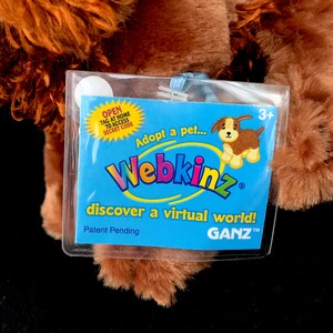 Op de afbeelding: Een bruin Webkinz pluche hondenspeelgoed met een blauw label waarop staat "Adopteer een huisdier... Webkinz ontdek een virtuele wereld!" en "GANZ" in een blauwe cirkel. Het label heeft ook een klein plaatje van een bruine en witte hond.