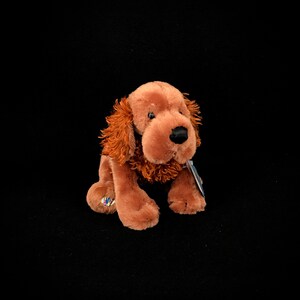 Op de afbeelding: Een bruine pluche hond met slappe oren en een bruine neus. De hond zit op een zwarte achtergrond.