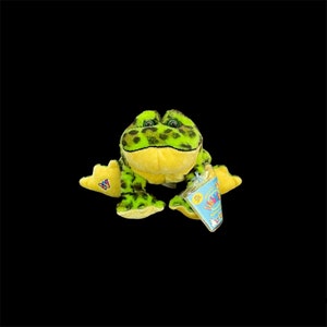 Pode incluir: Um brinquedo de pelúcia de sapo com um padrão de manchas verdes e pretas na cabeça e nas costas, e detalhes amarelos na barriga e nos pés. O brinquedo tem uma etiqueta anexada com um design azul e branco.