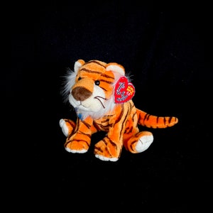 Puede incluir: Un peluche de tigre con rayas naranjas y negras, detalles blancos y una etiqueta roja en forma de corazón. El tigre tiene una melena blanca y una nariz marrón. El juguete está sobre una superficie negra.