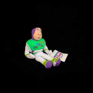 May include: A Buzz Lightyear plush toy from the Disney Store. The toy is wearing a green and white spacesuit with purple accents and a purple hood. A Disney Store tag is attached.