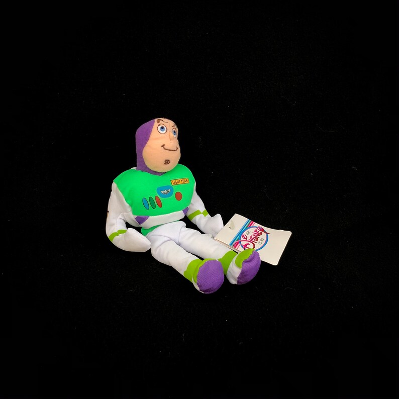 May include: A Buzz Lightyear plush toy from the Disney Store. The toy is wearing a green and white spacesuit with purple accents and a purple hood. A Disney Store tag is attached.