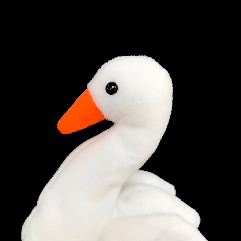 Op de afbeelding: Een witte pluche gans speelgoed met een oranje snavel en zwarte ogen, tegen een zwarte achtergrond. De nek van het knuffel is gebogen en het lichaam is gedeeltelijk zichtbaar. Kinderspeelgoed.