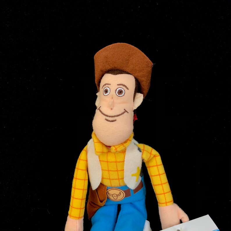 May include: A plush toy of the character Woody from the movie Toy Story. Woody wears a brown cowboy hat, a yellow plaid shirt, a white vest, blue pants, and a brown belt with a buckle. He has a sheriff's badge and a brown holster.