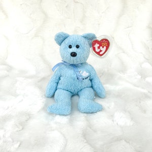 May include: A light blue Beanie Baby teddy bear with a white swan embroidered on its chest. The bear has a red Ty tag attached to its ear. The bear is sitting on a white fluffy surface.