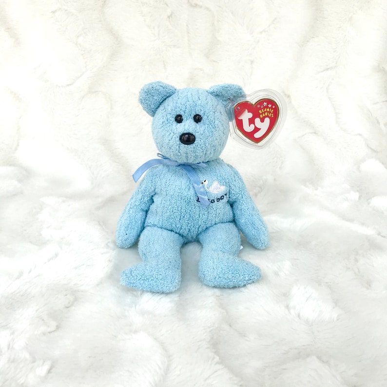 May include: A light blue Beanie Baby teddy bear with a white swan embroidered on its chest. The bear has a red Ty tag attached to its ear. The bear is sitting on a white fluffy surface.
