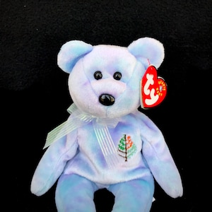 May include: A blue and purple tie-dye beanie baby bear with a black nose and brown eyes. The bear is wearing a white ribbon and has a small embroidered tree on its chest. The bear has a red heart-shaped tag with the words "Beanie Babies" and a snowflake.