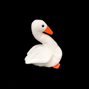 Op de afbeelding: Een witte pluche gans speelgoed met een oranje snavel en poten. De gans heeft zwarte ogen en staat tegen een zwarte achtergrond. Het speelgoed is zacht en pluizig en de gans staat in een gebogen positie.