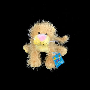May include: A plush lion with a pink nose and yellow fur. The lion is sitting with its paws tucked under its body. It has a Webkinz tag attached to its leg that says "Discover a virtual world".
