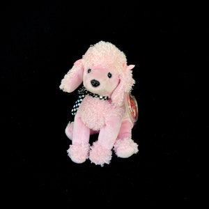 Puede incluir: Un peluche de caniche rosa con un lazo de lunares negros y blancos alrededor del cuello. El peluche tiene una etiqueta roja y blanca pegada en la espalda.