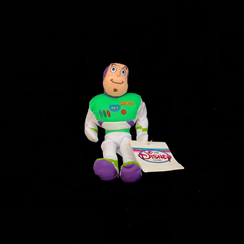 May include: A plush Buzz Lightyear doll from the Disney Store. The doll features a green and white spacesuit with purple accents, a smiling face, and a tag with the Disney Store logo. The doll is approximately 6 inches tall.