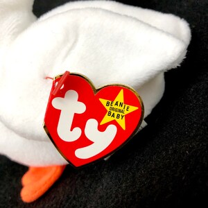 Op de afbeelding: Een witte pluche gans met oranje voeten. Een rood hartvormig label met het witte "ty" logo en "BEANIE ORIGINAL BABY" in gele tekst is aan het speelgoed bevestigd.