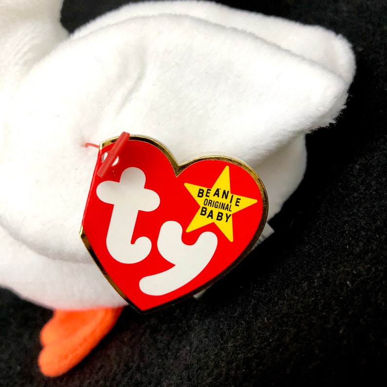 Op de afbeelding: Een witte pluche gans met oranje voeten. Een rood hartvormig label met het witte "ty" logo en "BEANIE ORIGINAL BABY" in gele tekst is aan het speelgoed bevestigd.