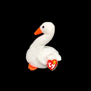 Op de afbeelding: Een witte pluche gans met een oranje snavel en poten. De gans heeft een lange, gebogen nek en een rood hartvormig label aan de zijkant. Op het label staat het woord "Ty".