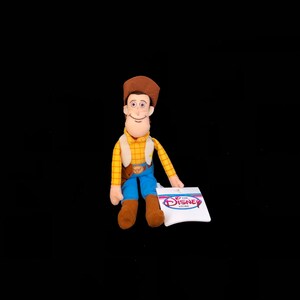 May include: A plush Woody doll from the Disney Store. The doll is wearing a brown cowboy hat, a yellow plaid shirt, a tan vest, blue pants, and brown boots. A white tag with "The Disney Store" logo is attached.