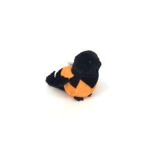 Puede incluir: Un juguete de peluche de un pájaro negro y naranja con detalles blancos. El pájaro está sentado sobre una superficie blanca.