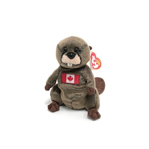 Può includere: Un castoro di peluche marrone e grigio con una bandiera canadese sul petto. Il castoro indossa un'etichetta rossa Ty Beanie Baby con il testo "Beanie Babies" e un logo "Ty" bianco.