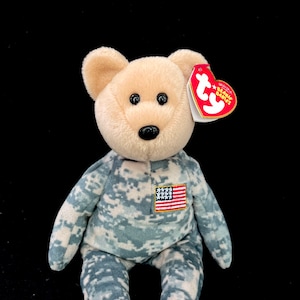 May include: A beige camouflage beanie baby bear with a red and white American flag patch on its chest. The bear is wearing a camouflage outfit and has a red "Ty" tag attached to its ear.