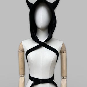 Puede incluir: Prenda negra con capucha y cuernos, usada como accesorio de disfraz. La capucha está atada al cuello y la prenda tiene un cinturón con detalles metálicos.