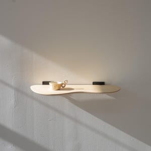 May include: A light-colored, wooden shelf with a unique, curved shape, mounted on a gray wall. A small, patterned teacup with a looped handle sits on the shelf. The shelf is supported by two black brackets.