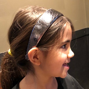 May include: A young girl with long brown hair wearing a black top and a silver snake-skin patterned headband.