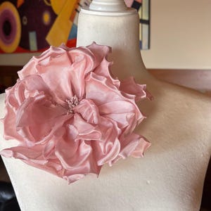 May include: A large, light pink fabric flower, possibly a brooch or decorative element, is displayed on a mannequin. The flower has multiple layers of ruffled petals and a textured center. The mannequin is white, and the background includes a colorful abstract painting.
