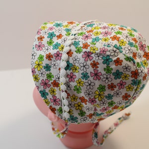 Puede incluir: Un gorro de beb&eacute; blanco con estampado floral en tonos rosa, amarillo, verde y naranja. El gorro tiene un ribete de encaje blanco alrededor del borde.
