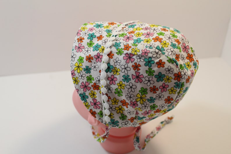 Puede incluir: Un gorro de beb&eacute; blanco con estampado floral en tonos rosa, amarillo, verde y naranja. El gorro tiene un ribete de encaje blanco alrededor del borde.
