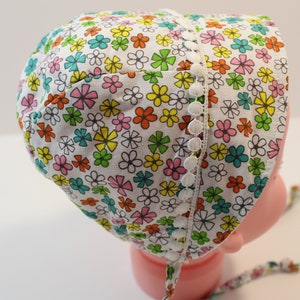 Puede incluir: Un gorro de beb&eacute; blanco con estampado floral en tonos rosa, naranja, amarillo, verde y azul. El gorro tiene un ribete de encaje blanco con pompones blancos.