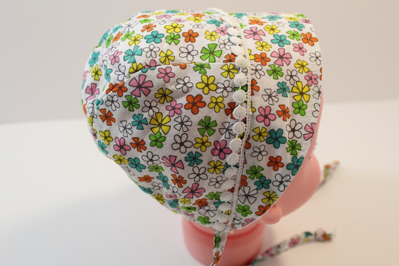 Puede incluir: Un gorro de beb&eacute; blanco con estampado floral en tonos rosa, naranja, amarillo, verde y azul. El gorro tiene un ribete de encaje blanco con pompones blancos.