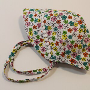 Puede incluir: Un sombrero de sol blanco con estampado floral en tonos rosa, amarillo, verde y azul. El sombrero tiene un ribete blanco con pompones blancos alrededor del borde.