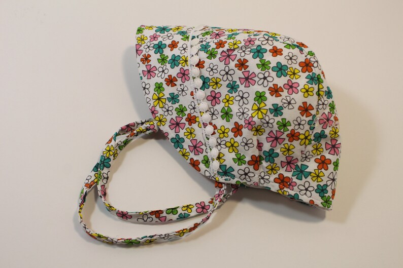 Puede incluir: Un sombrero de sol blanco con estampado floral en tonos rosa, amarillo, verde y azul. El sombrero tiene un ribete blanco con pompones blancos alrededor del borde.