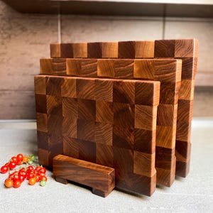 May include: Three wooden cutting boards with a checkered pattern, stacked on top of each other. The boards are made of dark wood and have a natural finish. The boards are sitting on a wooden stand.