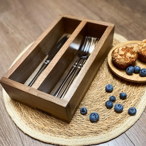 May include: A wooden cutlery organizer with three compartments, holding a knife and forks. Two muffins and blueberries sit on a small wooden plate. The organizer and plate rest on a woven placemat.
