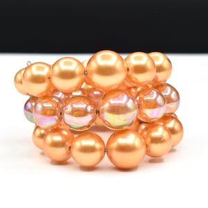 May include: A cluster bracelet featuring large, round, peach-colored beads. Some beads have an iridescent, multi-colored sheen. The bracelet is made of multiple strands of beads, creating a stacked effect. The beads are strung together with a visible string.