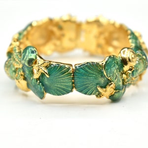 May include: Gold-toned hinged bracelet with a sea-themed design. The bracelet features teal-colored enamel shells and starfish accents. The intricate design is set against a bright white background, highlighting the jewelry's details.