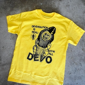 Op de afbeelding: Geel T-shirt met een zwarte grafische print van een persoon met een helm en een microfoon. De tekst "DE-EVOLUTION IS REAL! WE'RE ALL DEVO" is op het T-shirt gedrukt.