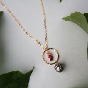 May include: A gold chain necklace with a small circle pendant. The pendant has a cluster of pink and red gemstones hanging from it. A large, dark gray pearl hangs from the bottom of the circle.