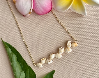 May include: A gold chain necklace with a row of small white seashells. The necklace is laying on a beige fabric background with pink and yellow flowers.