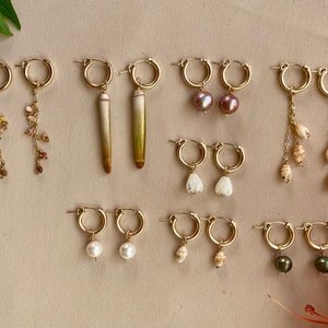 May include: A collection of gold hoop earrings with various charms, including seashells, pearls, and flowers. The earrings are displayed on a beige surface.