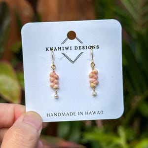 May include: Dangle earrings featuring small, braided pink and cream-colored shells suspended from gold-tone hooks, each accented with a tiny white pearl at the base. The earrings are displayed on a white card labeled KWAHIWI DESIGNS.