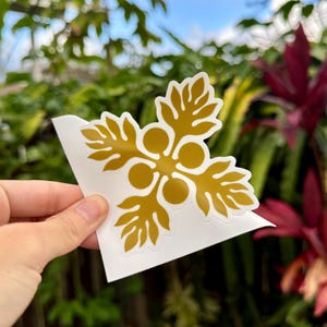 May include: A gold-colored vinyl sticker with a stylized flower design. The sticker is on a white background and is being held by a hand.